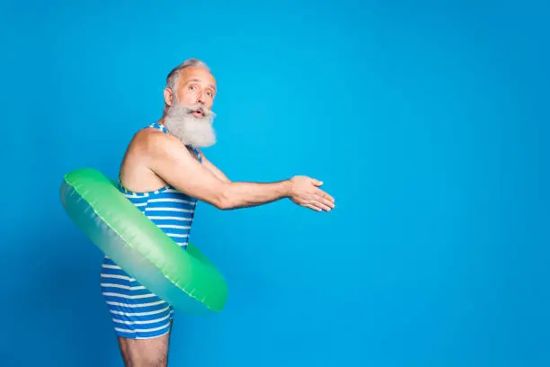 Profile side photo of impressed old man jumping with rubber round circle wearing striped swim wear isolated over blue background