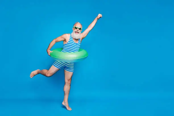 Photo of Full length photo of excited retired pensioner with grey hair raising his fist holding toy circle wearing striped swimwear glassses isolated over blue background