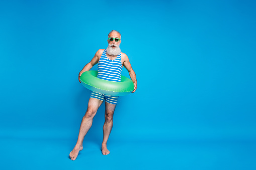 Full size photo of impressed man in eyeglasses eyewear wearing striped bathing costume hold lifesaver trying ocean water isolated over blue background