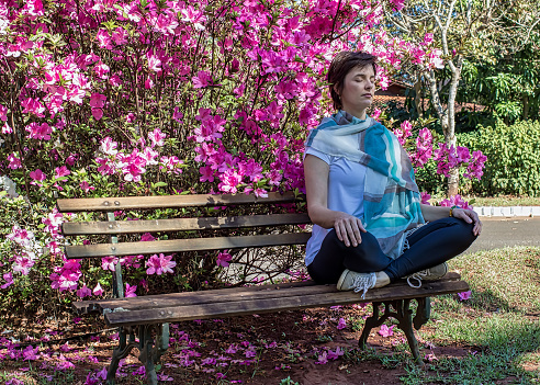 Model 40 years old woman sitting on wooden bench and meditating in the park in front of an azaleas bush - 40-year-old or middle-aged woman meditating in park