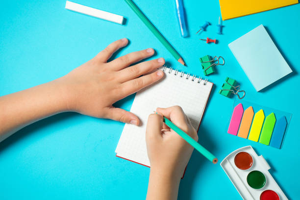 istock children's hand writes in a notebook in composition with watercolor, pencil, notebooke, ruler, eraser. Isometric concept on blue background. Pop art. School supplies. overhead. back to school concept 1169155789