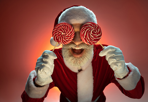 Cheerful mature Santa Claus standing in costume and covered face holding lollipops