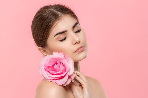 Close-up portrait of a beautiful young girl holding pink rose flower close to face isolated over pink background. Concept of beauty and health care.