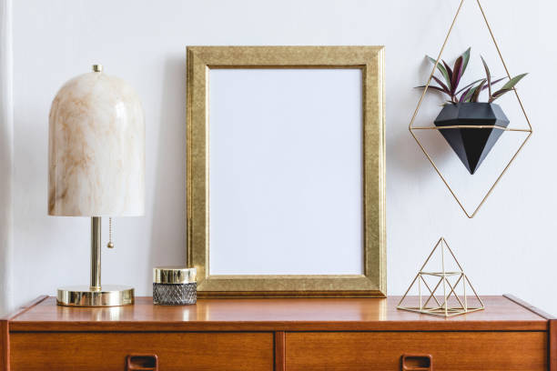 istock Stylish composition of open space interior with gold mock up frame,retro wooden commode, marble table lamp, gold pyramid, plants and elegant accessories. Modern design interior. Template. Ready to use 1169161871