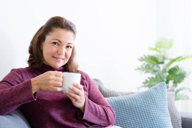 istock Middle aged lady sitting on sofa holding coffee or tea mug 1169176040