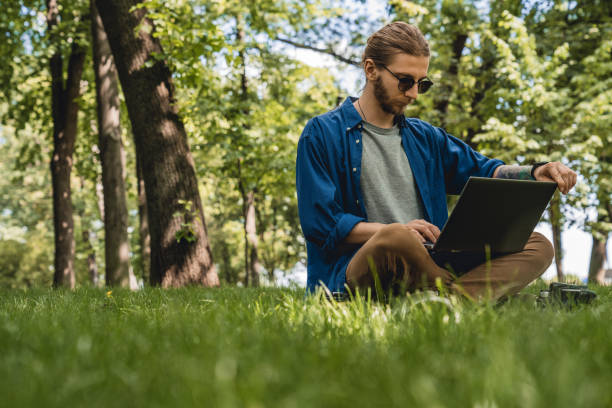 istock Young man sitting on grass at park working on laptop 1169196046