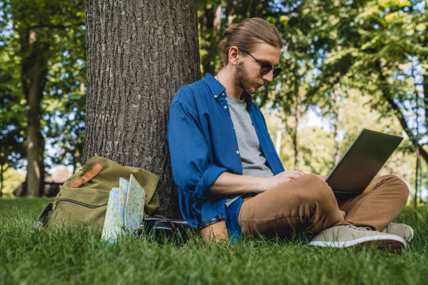 istock Male wearing sunglasses using laptop while sitting under a tree at park 1169196105
