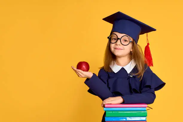 Photo of Cheerful schoolgirl in graduation outfit carrying apple while standing beside pile of textbooks