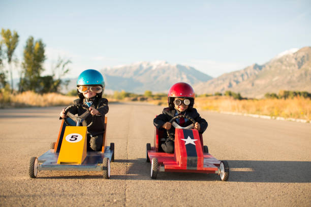 Young Business Boys Racing Toy Cars Two young boys dressed as businessmen wearing racing goggles and helmets sit in their toy go carts and cars on a rural road in Utah, USA. One car is yellow and the other is red and they love a healthy business competition. rivalry stock pictures, royalty-free photos & images