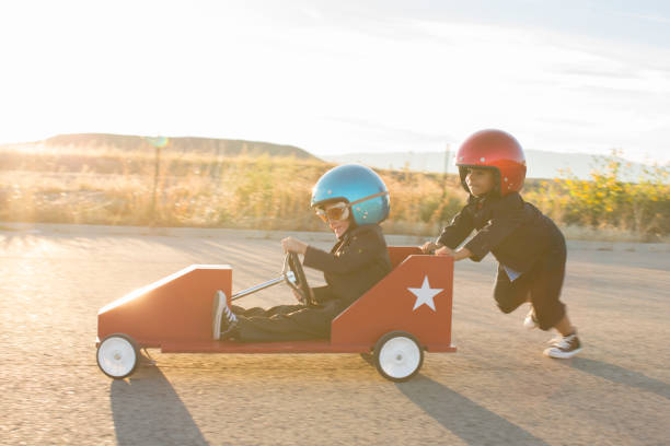Young Business Boys Racing a Toy Car Two young boys dressed as businessmen wearing racing goggles and helmets race their toy go cart and car on a rural road in Utah, USA. One boy pushes the other business boy while working together towards a finish line. auto racing photos stock pictures, royalty-free photos & images