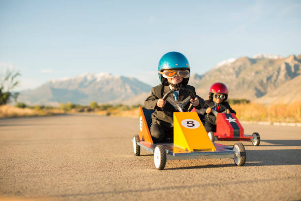 Young Business Boys Racing Toy Cars Two young boys dressed as businessmen wearing racing goggles and helmets sit in their toy go carts and cars  racing on a rural road in Utah, USA. One car is yellow and the other is red and they love a healthy business competition. motorsport photos stock pictures, royalty-free photos & images
