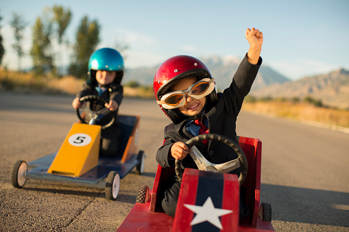 A young boy dressed as a businessman wearing racing goggles and helmet raises his arm in victory as he has won the business race and earned glory. He is racing his go cart and favorite car on a rural road in Utah, USA.