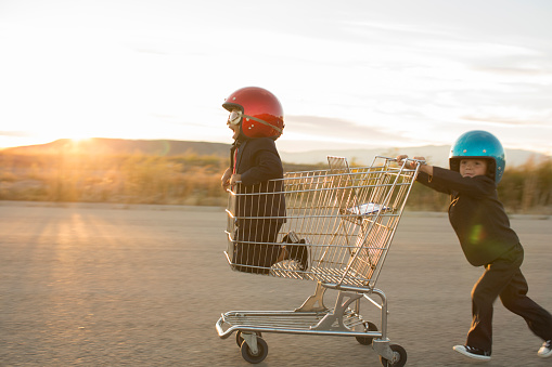 Two young boys dressed as businessmen wearing racing goggles and helmets race a shopping cart on a rural road in Utah, USA. One boy pushes the other business boy while working together towards a finish line.