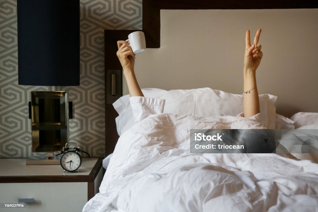 Unrecognizable  woman waking up in the morning in the bed, hiding under the blanket, holding a cup of coffee and showing the peace sign Bed - Furniture Stock Photo