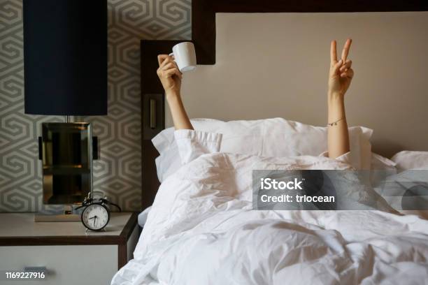 Unrecognizable Woman Waking Up In The Morning In The Bed Hiding Under The Blanket Holding A Cup Of Coffee And Showing The Peace Sign Stock Photo - Download Image Now