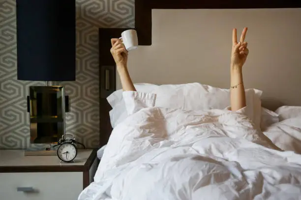 Unrecognizable  woman waking up in the morning in the bed, hiding under the blanket, holding a cup of coffee and showing the peace sign