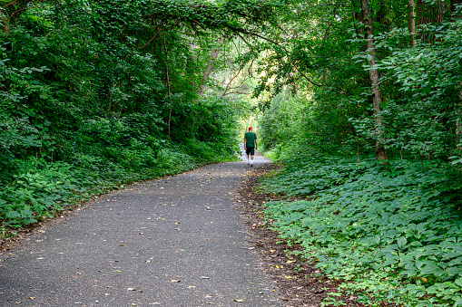 Wide angle distant image of one older aged Latino man walking down heavily wooded pathway.  At the end of pathway the light silhouettes the man.\n\nTaken in Minneapolis. Minnesota, USA