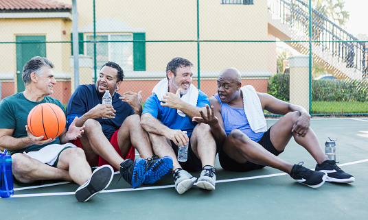 A group of four multi-ethnic mature and senior men in their 40s, 50s and 60s, taking a break after playing basketball. They are sitting on the outdoor court with water bottles and towels, relaxing, and talking.
