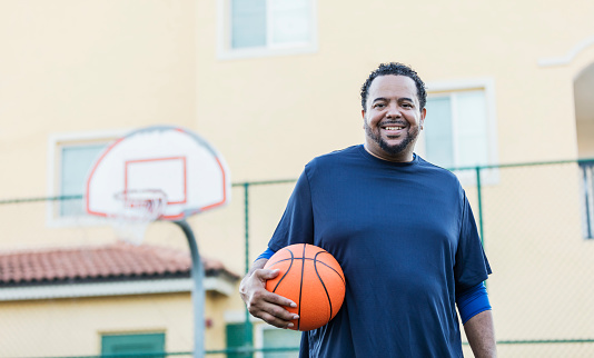 A mature African-American man in his 40s standing on an outdoor basketball court, holding a ball, smiling at the camera.