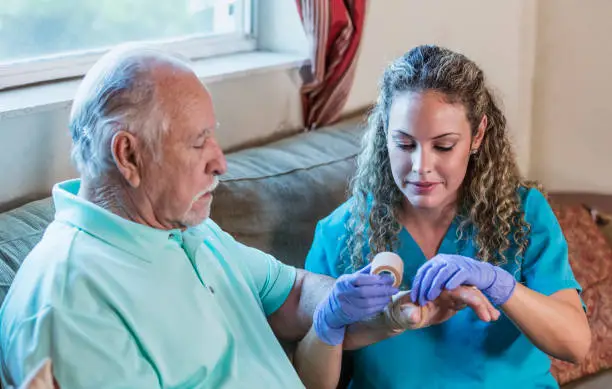 A mid adult Hispanic woman in her 30s working as a home health aide or nurse, helping an senior man in his 80s, wrapping his hand in a bandage.