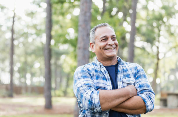 Mature Hispanic man wearing plaid shirt A mature Hispanic man in his 50s wearing a plaid shirt, standing in a park, smiling with his arms crossed. checked pattern photos stock pictures, royalty-free photos & images