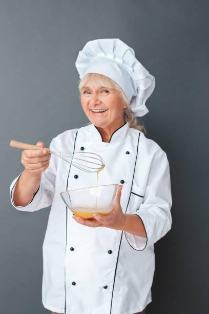 istock Senior woman chef studio standing isolated on gray holding whisk mixing eggs looking camera cheerful 1169247479