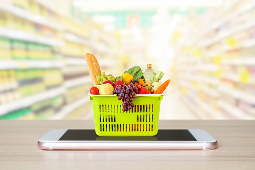 Fresh food and vegetables in shopping basket on mobile smartphone on wood table with supermarket aisle blurred background grocery online concept