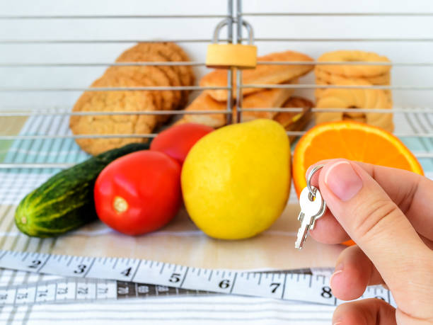 Woman hand holds the key of cell with cookies. Cookies are in the cage locked with padlock. Vegetables, fruits and measuring tape with centimeters and inches are in the foreground. Healthy eating and ban on junk food concept. Woman hand holds the key of cell with cookies. Cookies are in the cage locked with padlock. Vegetables, fruits and measuring tape with centimeters and inches are in the foreground. Healthy eating and ban on junk food concept. Front view. tomato cages stock pictures, royalty-free photos & images