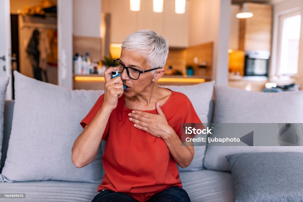 Asthmatic woman using an inhaler Senior gray hair woman wearing eyeglasses, feeling unwell and coughing as symptom for cold or bronchitis. Healthcare concept.Front view of mature Woman using asthma inhaler while sitting on sofa ta home during the day. Short hair woman with flu inhalation. Photo of mature woman using asthma inhaler. Asthmatic Stock Photo