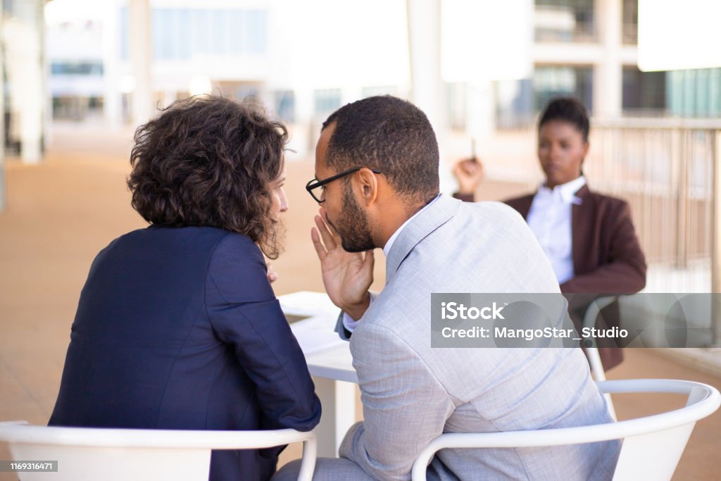 Employees gossiping about young female colleague Employees gossiping about young female colleague. Business man and woman whispering, African American employee sitting in background. Office rumors concept Gossip Stock Photo