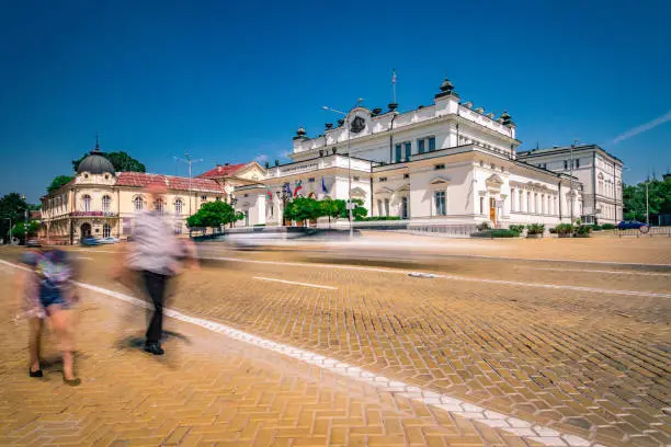 Panoramic long exposure day time photo from Tsar Osvoboditel square in Sofia city centre in Bulgaria, Eastern Europe. Unrecognisable blurred real people in the foreground and the iconic architecture of the National Assembly of the Republic of Bulgaria (House of Parliament) and the building of Library of Bulgarian Academy of Sciences in the far background, unrecognisable people, etc. Dragged shutter technique used to capture the blurred headlight/taillights from the road traffic and unrecognisable commuters. Shot on Canon full frame EOS R system for premium quality.