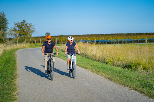 real people happy active seniors couple cycling together on electric bicycles through rural summer landscape on cycling path small asphalt road