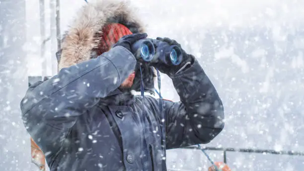 Photo of Polar Scientist and Adventurer in Warm Jacket Standing on Ship and Looking through Binoculars. Polar Research Exploration During Winter Storm.