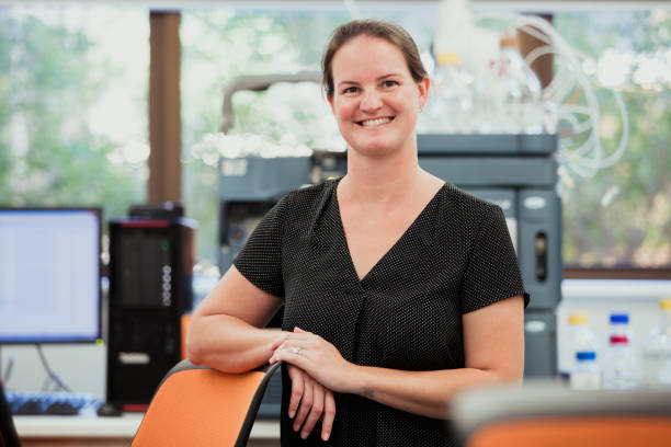 University Researcher At Work Female scientist looking at the camera leaning on a chair while smiling. She is a biochemist working in a university doing phenotype research. biochemist stock pictures, royalty-free photos & images