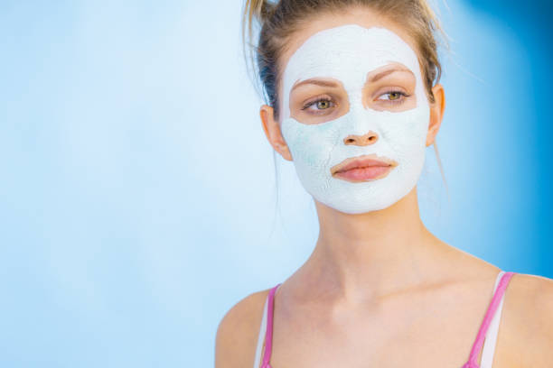 Young woman with white dry mud mask on face, against blue. Teen girl taking care of oily skin, cleaning the pores. Beauty treatment. Skincare.