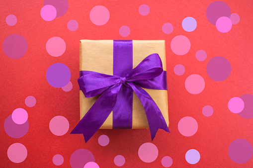 Giftbox tied with purple color ribbon on dark red background with beautiful confetti. Flat lay style.