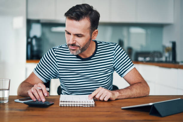 istock Smiling man managing home budget with calculator and notebook while sitting at dining table 1169342851