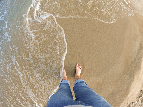 Top view of man's bare feet while standing on a sandy beach with sea surfs splashing. Seen from the first person perspective, POV.
