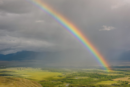 Amazing bright colorful rainbow over the mountains, a valley with a winding river and forest against a stormy sky with clouds and rain. Altai, Russia.