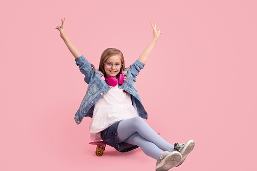 Sweet kid in stylish denim outfit cheerfully smiling and gesturing V sign with raised hands while sitting on modern longboard against pink background