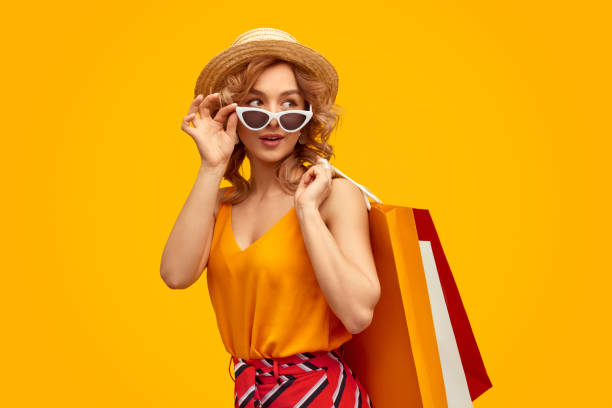 Stylish shopaholic with purchases Trendy young woman with paper bags adjusting sunglasses and looking away over shoulder during shopping against bright yellow background bag photos stock pictures, royalty-free photos & images