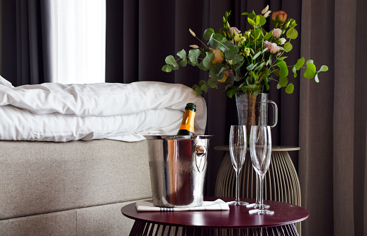 Romantic setting in the bedroom with open champagne bottle and empty champagne glasses on table in the bedroom with fresh flower bouquet in a vase in the background
