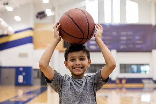 In the gym after school, the elementary age boy goofs off with a basketball for the camera.