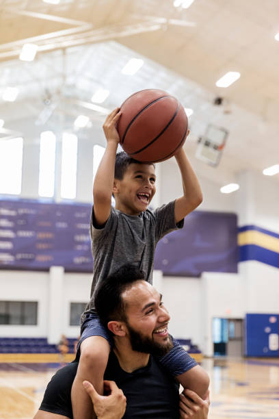 With nephew on uncle's shoulders, both have fun shooting baskets The mid adult uncle puts his young nephew on his shoulders and both have fun shooting baskets at the gym. youth basketball indoor stock pictures, royalty-free photos & images