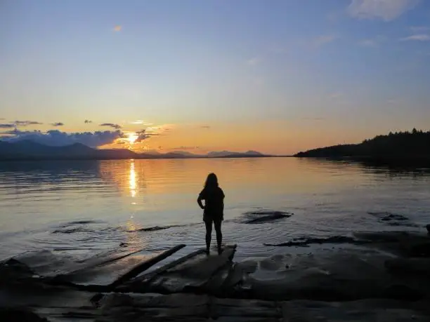 Photo of A young female traveller standing at the edge of the shoreline staring at the open ocean.  The sunset has almost set and is reflecting off of the ocean.  It is a beautiful evening.  True wanderlust.