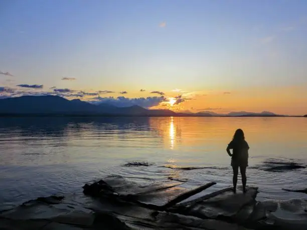 Photo of A young female traveller standing at the edge of the shoreline staring at the open ocean.  The sunset has almost set and is reflecting off of the ocean.  It is a beautiful evening.  True wanderlust.