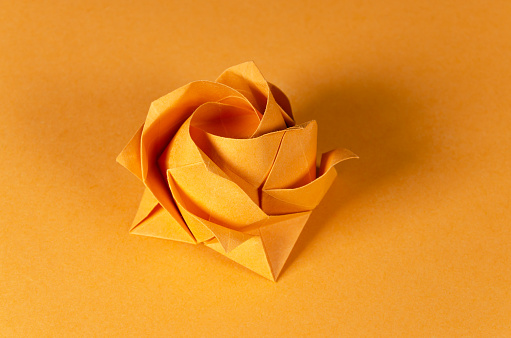 Orange origami rose on orange background. Japanese art of paper folding. Flat square sheet of paper transferred into a finished sculpture through folding and sculpting. Close up. Macro photo.