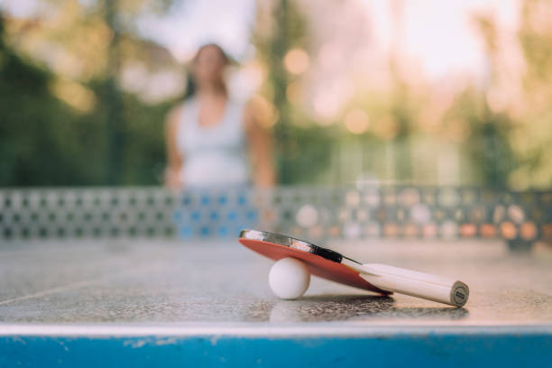primo piano della racchetta da ping pong e palla sul tavolo con donna in piedi sullo sfondo - table tennis table tennis racket racket sport ball foto e immagini stock