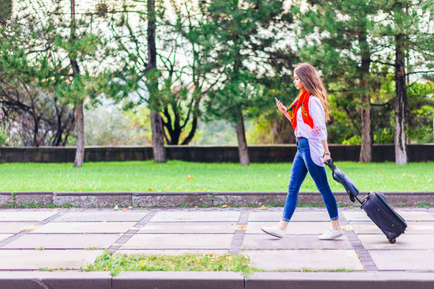 istock Young woman using her smart phone  and pulling a small wheeled luggage with a briefcase on it 1169535600
