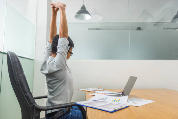 Tired Asian employee stretching in front of computer at office stock photo